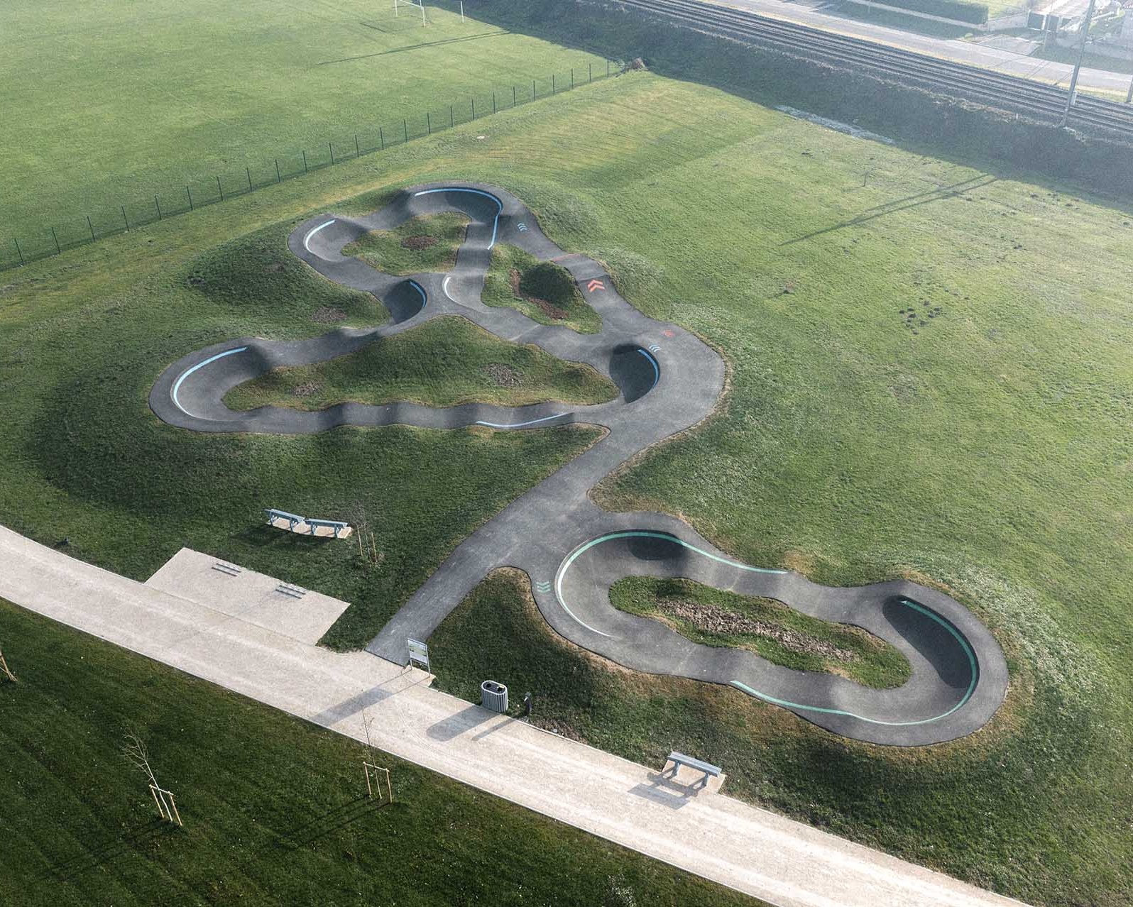 Luftaufnahme eines modernen, geschwungenen Asphalt-Pumptracks auf einer grünen Wiese neben Bahngleisen.